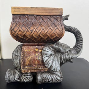 May include: A decorative elephant figurine with a woven basket on its back. The elephant is gray with gold accents on its saddle. The basket is brown and has a woven texture. The elephant's trunk is curled upwards.