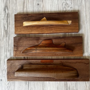 May include: Three wooden submarine sculptures, each mounted on a rectangular walnut base. The submarines are carved with varying designs and colors, showcasing the natural wood grain. The top submarine is light-colored, the middle one has a darker stripe, and the bottom one is a darker wood.