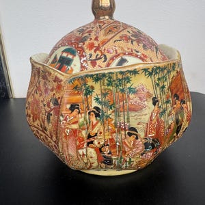 May include: A decorative, lidded ceramic jar with a gold-colored knob. The jar features a detailed, hand-painted design depicting figures in traditional Japanese attire, set against a backdrop of bamboo and floral patterns. The color palette includes gold, red, and cream.