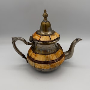 May include: A metallic teapot featuring a curved handle and spout, with a lid topped by a finial. The body and lid are decorated with inlaid rectangular pieces of light tan, marbled material arranged in bands, separated by thin copper-toned wire.