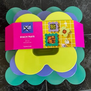 May include: A set of three colorful snack trays in shades of green, yellow, and purple. The trays have a scalloped edge design. A pink band with the text "SNACK TRAYS" and the brand name is wrapped around the trays.