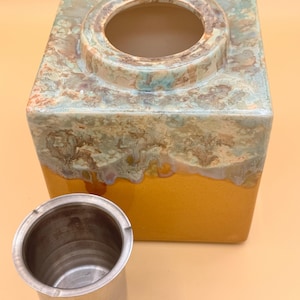 May include: A square ceramic tea caddy with a circular opening, featuring a two-tone design. The top half has a mottled turquoise and brown glaze, while the bottom is a solid gold color. A small, stainless steel tea infuser is next to the caddy.