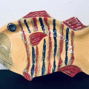 May include: A ceramic fish-shaped serving dish with a yellow base and red and blue vertical stripes. The dish has red fins and a blue eye. The dish is likely handmade and has a unique, artistic design.