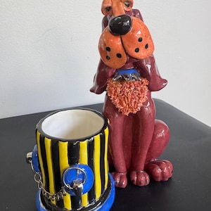 May include: A ceramic figurine of a maroon dog with long ears and a black nose, positioned next to a cylindrical ceramic container painted with vertical yellow and black stripes, featuring a blue base and a small silver chain detail.