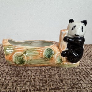 May include: A ceramic planter shaped like a log with a panda bear clinging to a tree trunk. The planter is painted with shades of brown, green, and orange. The panda has black and white fur.