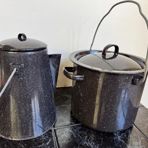 May include: Two vintage black enamelware pots with white speckles. One is a coffee pot with a lid and handle, the other is a pot with a lid and a wire handle. Both are on a black tiled surface.