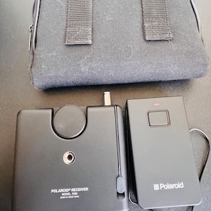 May include: A black Polaroid Receiver Model 7030 and a black Polaroid remote control are displayed next to a black carrying case. The receiver has a circular dial and a jack. The remote has a red light and the Polaroid logo.