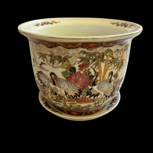 May include: Cream-colored ceramic planter with a matching saucer. The planter features a detailed Asian-inspired design with cranes, bamboo, and floral elements in shades of red, gold, and green. The rim has a gold trim.