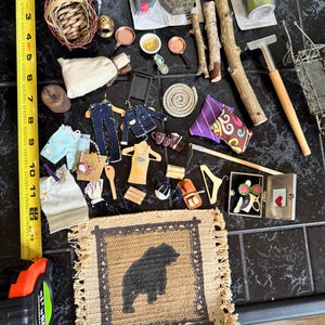 May include: A collection of miniature craft supplies and decorative items, including woven baskets, tiny clothing, metal lanterns, wooden sticks, and a small rug with a bear silhouette. A measuring tape is also visible.
