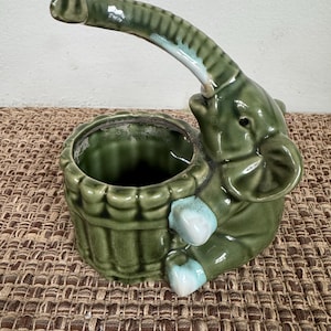 May include: A green ceramic elephant planter with a raised trunk forming a handle. The planter has a round opening and is decorated with light blue accents on the feet and trunk. The elephant is sitting next to a round planter.