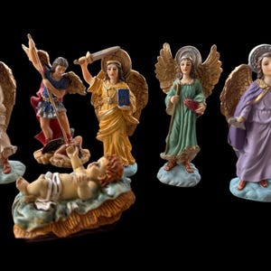May include: A collection of detailed angel figurines with gold-colored wings, halos, and flowing robes. One figure holds a sword, another a book, and another a fish. A baby figurine rests in a manger. The figurines are on a black background.