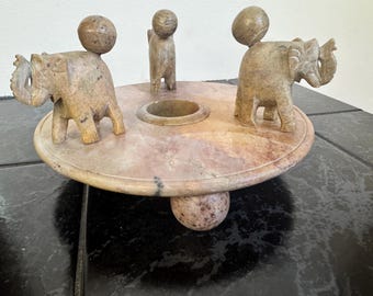May include: A decorative, handcrafted stone centerpiece featuring two elephants and a small figure on a circular base. The elephants have a ball on their backs. The base is supported by a large ball. The color is a mix of beige and pink.