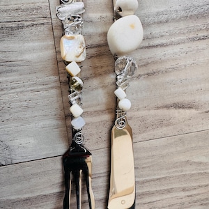 May include: Two handcrafted serving utensils: a fork and a butter knife. The handles are adorned with a variety of clear and white beads, stones, and a mother-of-pearl square. The utensils are made of silver-colored metal.