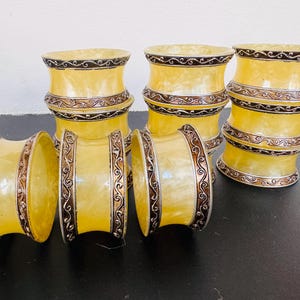May include: Set of six decorative yellow napkin rings. Each ring features a metallic silver and brown ornate design. The napkin rings are stacked in groups of three and arranged on a dark surface.