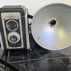 May include: Vintage Kodak Duaflex II camera with a silver flash attachment. The camera is black with silver accents and features a lens with settings. The flash is a large, round reflector. A black strap and cord are attached.