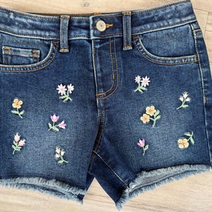 May include: Dark blue denim shorts with embroidered flowers in pink, yellow, and green. The shorts have a frayed hem.