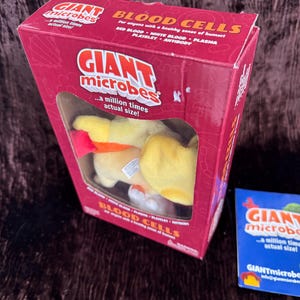 May include: A red and white box with a plush toy inside. The box has the words "Giant Microbes" and "Blood Cells" on it. The toy is a yellow and orange plush blood cell.