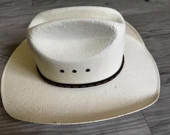 May include: A white cowboy hat with a wide brim and a brown band. The hat has a textured surface and three small black dots on the front. The hat is made of straw.