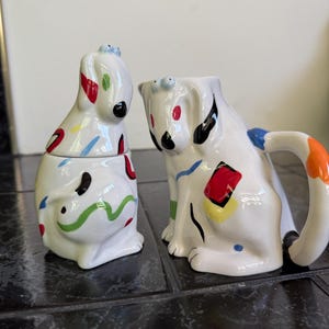 May include: A white ceramic creamer and a matching lidded sugar bowl, both shaped like stylized dogs. The pieces feature hand-painted abstract accents in red, blue, yellow, green, and black, with prominent eyes and ears on each dog figure.