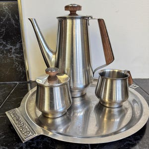 May include: A vintage stainless steel coffee set with a tray. The set includes a coffee pot, creamer, sugar bowl, and a round tray with decorative handles. The coffee pot and sugar bowl have wooden accents.
