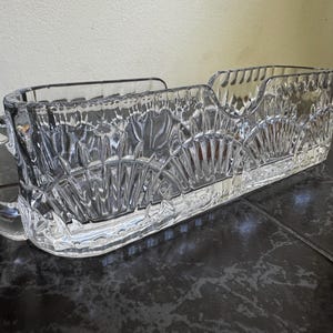 May include: A clear, rectangular crystal serving dish with a handle on one end. The dish features a cut-glass design with a series of vertical and fan-shaped patterns. The handle is rounded and smooth, and the dish sits on a dark surface.