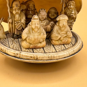 May include: A detailed ivory-colored sculpture featuring multiple seated figures and a dragon, set within a shell-shaped structure. The figures are intricately carved, depicting various characters. The sculpture is placed on a decorative base.