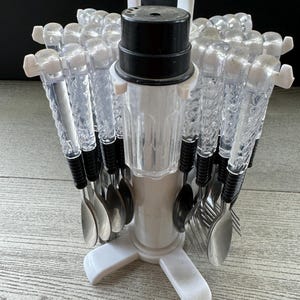 May include: A white plastic cutlery holder with a black top and a clear plastic container. The holder has a three-pronged base and holds a variety of spoons and forks.