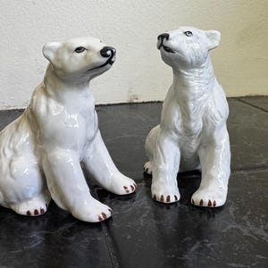 May include: Two white ceramic polar bear figurines. One bear is seated with its head turned to the side, and the other is sitting upright, looking up. Both have black noses and brown paws.