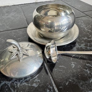May include: A silver-toned serving set, including a round bowl on a matching plate, a ladle, and a lidded dish with a tomato-shaped handle. The set is displayed on a black and gray tiled surface.