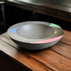 May include: A gray, round bowl with a wide rim. The rim is decorated with geometric shapes in pink, green, blue, and yellow. The bowl is sitting on a wooden surface. This decorative bowl is suitable for serving food or as a decorative item.