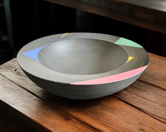 May include: A gray, round bowl with a wide rim. The rim is decorated with geometric shapes in pink, green, blue, and yellow. The bowl is sitting on a wooden surface. This decorative bowl is suitable for serving food or as a decorative item.