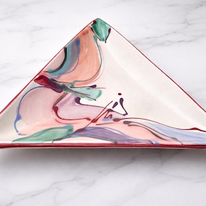 May include: Triangular ceramic plate with a white base and a colorful abstract design. The design features flowing lines and shapes in shades of pink, purple, green, and red. The rim is a deep red.
