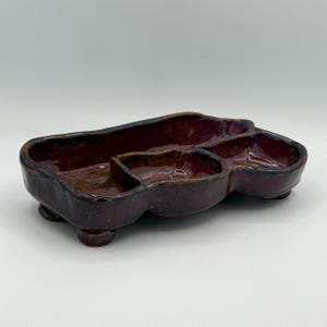 May include: A rectangular ceramic serving dish with a glossy dark burgundy glaze and an irregular, organic shape. The dish features three distinct interior compartments separated by low walls and is supported by four small, rounded ceramic feet.