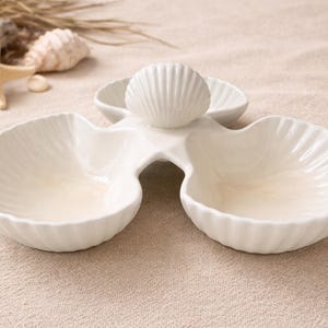 May include: A white ceramic serving dish with a seashell design. The dish has three sections, each shaped like a seashell. A starfish and other seashells are in the background. The dish is ideal for serving snacks or appetizers.