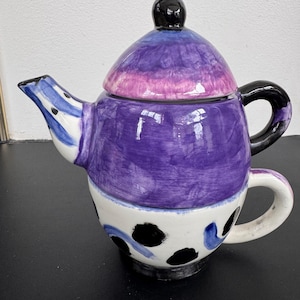 May include: A whimsical, hand-painted ceramic teapot with a matching teacup base. The teapot features a purple body, a black handle, and a lid with a black knob. The teacup base is white with black polka dots and blue accents.