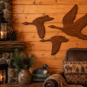 May include: Three brown wooden wall art pieces shaped like flying geese with detailed wing carvings. The geese are arranged in a flock formation on a horizontal wood plank wall, showcasing textured feathers and distinct silhouettes in flight.