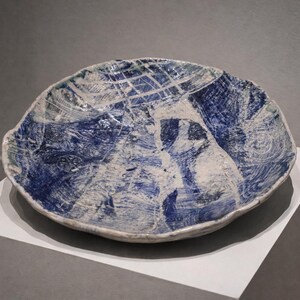 May include: A handcrafted ceramic bowl with a unique, organic shape. The bowl features a striking blue and white abstract design, with a textured surface. The bowl is displayed on a white pedestal, showcasing its artistic qualities.