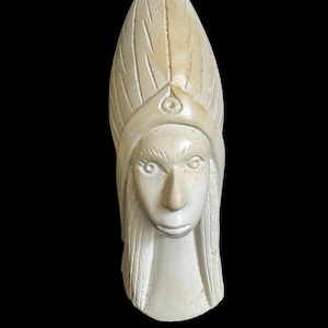 May include: Ivory-colored carved sculpture of a stylized human head with long hair and a tall headdress. The face has defined features, including eyes, a nose, and a mouth. The headdress has a central design and vertical lines.