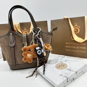 May include: A brown suede handbag with a top handle and decorative charms, including a teddy bear, camera, and tiger. The bag has a leather strap and is next to a marble-patterned box and shopping bags with "NEW COLLECTION" printed on them.