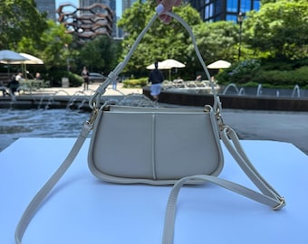 May include: A cream-coloured shoulder bag with a long, adjustable strap. The bag has a rectangular shape with rounded corners and gold-coloured hardware. The background shows a park with trees and buildings.