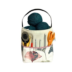 May include: A white canvas tote bag with a floral print and a pink flower design. The bag has a black handle and is filled with balls of yarn, knitting needles, and a pair of scissors.