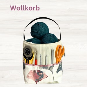 May include: A white fabric storage basket with a black handle, filled with green yarn balls and various knitting needles. The basket has a floral pattern and the text "Wollkorb" is printed on the top.