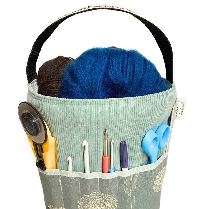 Puede incluir: Una bolsa de tela azul claro con un asa negra y un patr&oacute;n de dientes de le&oacute;n blancos. La bolsa est&aacute; llena de hilo, agujas de crochet, tijeras y una aguja de lana. La bolsa tiene una etiqueta que dice "Crochet hecho a mano".