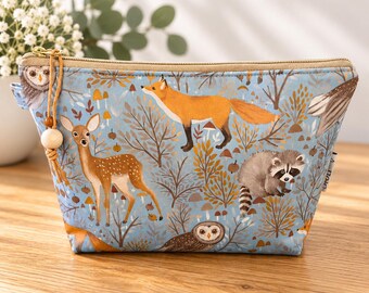 May include: A light blue zippered pouch featuring a woodland pattern with a deer, a fox, a raccoon, and owls among trees and mushrooms. The pouch has a tan zipper and a leather cord pull with a wooden bead attached to the side zipper pull.