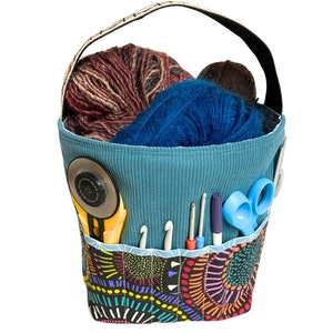 May include: A blue fabric tote bag with a colorful patterned bottom. The bag is filled with yarn, scissors, and other craft supplies.