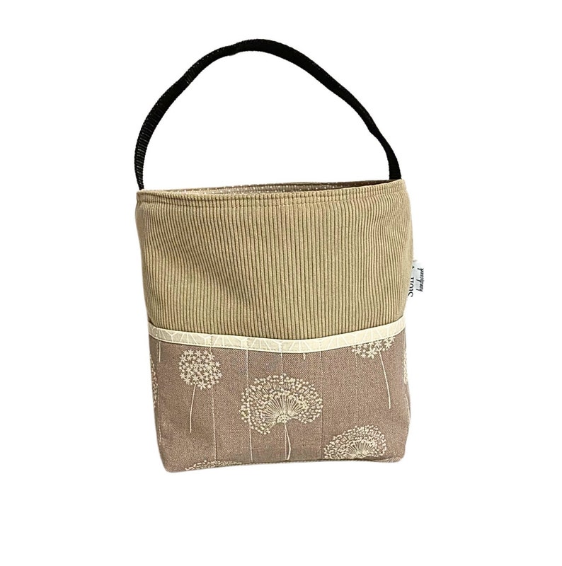 May include: A brown canvas tote bag with a black handle and a white dandelion pattern. The bag has a white trim and a brown corduroy top.