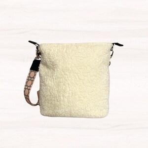 May include: Cream-colored, fluffy crossbody bag with a patterned strap. The bag has a soft, textured exterior and a black strap attachment. The patterned strap features a pink and white geometric design. The bag is designed to be worn over the shoulder.