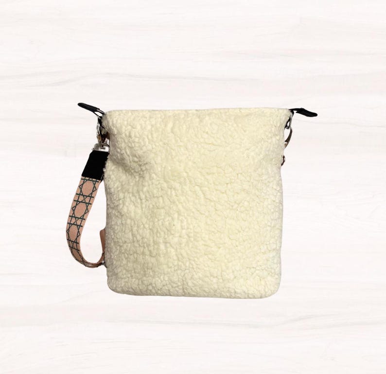 May include: Cream-colored, fluffy crossbody bag with a patterned strap. The bag has a soft, textured exterior and a black strap attachment. The patterned strap features a pink and white geometric design. The bag is designed to be worn over the shoulder.
