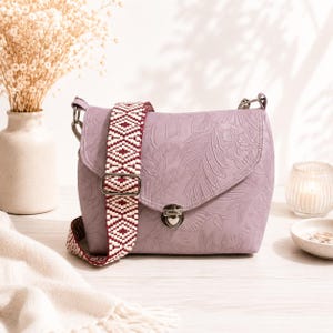 May include: A lilac crossbody bag with a patterned strap. The bag has a floral embossed design and a silver clasp. The strap features a woven pattern of white and burgundy diamonds. The bag is on a white surface.