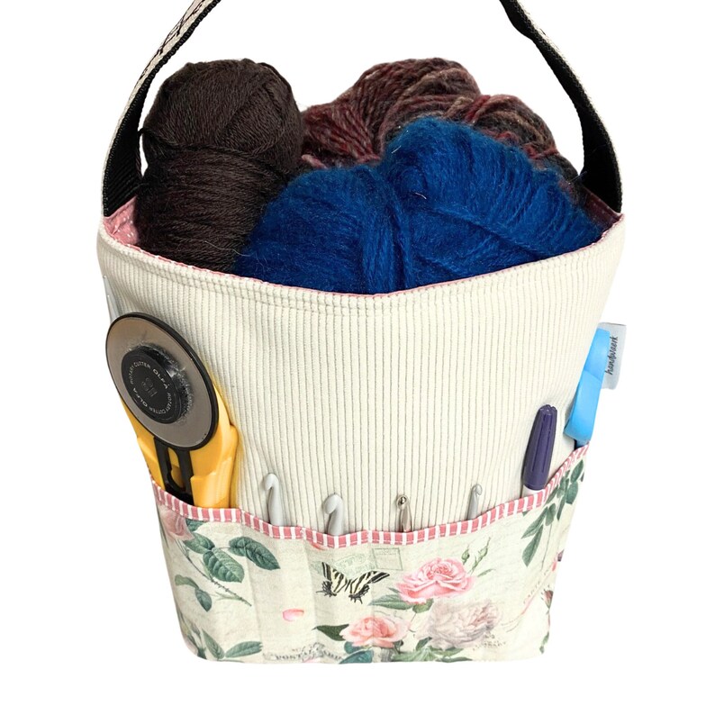 May include: A white canvas tote bag with a floral print and a pink trim. The bag is filled with yarn in brown, blue, and purple. The bag also has a yellow yarn cutter, a blue pen, and a blue and white sticker that says "Addict".